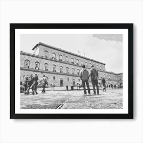 Exploring the Historic Building Palazzo Pitti. The image depicts a group of people walking and standing in front of a large, historic building with arched windows and a detailed facade. The scene is set in a spacious courtyard with a patterned stone pavement. The building's architecture and the presence of people suggest it is a place of interest, possibly a museum or a significant public building. Art Print