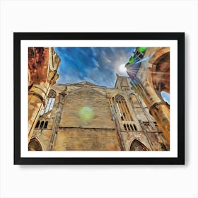 Stone Archway and Tower in Narbonne. The image captures a low-angle view of a weathered stone building with a prominent archway and a tower. The sun shines brightly in the blue sky, casting a warm glow on the aged stonework. The archway leads to a shadowy interior, while the tower stands tall against the clouds. 1 Art Print