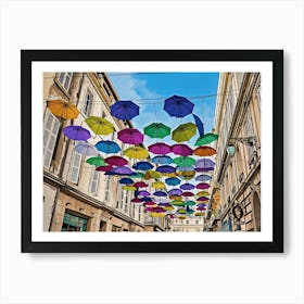 Colorful Umbrellas Street Art. A vibrant street scene features a multitude of colorful umbrellas suspended overhead, creating a whimsical and cheerful atmosphere. The umbrellas, in various shades of blue, green, purple, yellow, and pink, are strung across a narrow street lined with beige buildings. The buildings have white shutters and windows, adding to the European charm of the scene. Art Print