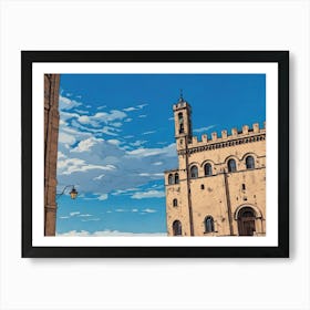 Palazzo dei Consoli (Palace of the Consuls) in Gubbio. A beautifully preserved medieval-style building with intricate stonework and arched windows under a vibrant blue sky. Its towering presence, complete with a clock tower, dominates a sunlit square where visitors stroll and admire the architecture. The scene captures the charm and history of a European town center. 1 Art Print