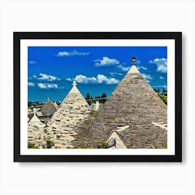 Trulli Houses in Italy. The image showcases a picturesque view of a traditional Italian village, featuring a cluster of Trulli houses with their distinctive conical roofs. The houses are constructed from white stone and have a rustic charm, with some roofs adorned with intricate patterns. The scene is bathed in warm sunlight, with a clear blue sky and fluffy white clouds overhead. 1 Art Print