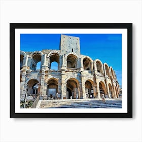 Roman Amphitheater in Arles. A majestic Roman amphitheater stands tall against a clear blue sky. The ancient structure is made of weathered stone, with numerous arches and a central tower. The amphitheater is surrounded by a set of stone steps leading up to the entrance. A few people are walking around the area, adding a sense of scale to the impressive monument. Art Print