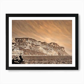 Cliffside View of Cap Canaille. The image captures a panoramic view of a rugged cliff face, its rocky surface bathed in warm, sepia tones. The cliff rises dramatically from the calm, blue waters below, creating a sense of grandeur and natural beauty. Two figures stand on a rocky outcrop at the edge of the cliff, their silhouettes barely visible against the vastness of the landscape. Art Print