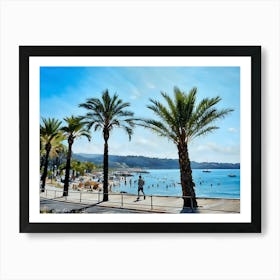 Palm Trees Beach Scene in French Riviera. A picturesque beach scene unfolds with a row of palm trees framing the view. The azure sky stretches above, while the clear blue water laps at the shore. A man in blue shorts jogs along the boardwalk, passing by a lively crowd of people enjoying the sun and sand. Colorful umbrellas dot the beach, adding a splash of vibrancy to the scene. Art Print