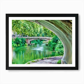 Tranquil Pathway Canal de Brienne Toulouse France. This image captures a tranquil riverside scene viewed from beneath a bridge. The lush greenery of the trees and bushes reflects beautifully on the calm, green-tinted water. In the background, another stone bridge spans the river, adding to the picturesque and serene atmosphere of the setting. Art Print