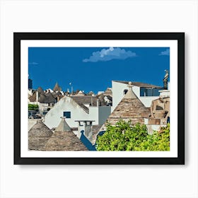 Trulli Rooftops in Italy. The image showcases a picturesque view of traditional Trulli houses in a small Italian town. The Trulli, characterized by their distinctive conical roofs made of dry-stone, are painted in a warm, earthy palette of browns and tans. The whitewashed walls of the houses contrast beautifully with the vibrant green foliage of a tree in the foreground. Art Print