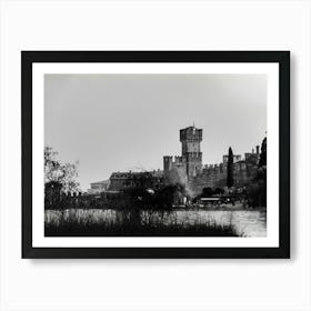 Medieval Castle by the Lake Garda Castello Scaligero Sirmione. A black and white photograph captures a majestic medieval castle standing tall against a cloudy sky. The castle, with its imposing stone walls and a prominent tower, dominates the scene. The foreground features a tranquil lake reflecting the castle's silhouette, with tall grasses and trees framing the view. Art Print