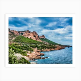 Coastal Cliffs and Blue Sea in Esterel France. The image captures a breathtaking coastal landscape with a dramatic interplay of land and sea. Rugged, reddish-brown cliffs rise from the shore, covered in lush green vegetation. The cliffs are punctuated by rocky outcroppings and a winding road that snakes along the hillside. Art Print