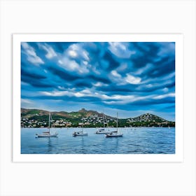 Sailboats at Anchor in Esterel France. The image captures a serene scene of several sailboats anchored in a calm bay. The boats are white with their masts raised, suggesting a peaceful and tranquil atmosphere. The water is a deep blue, reflecting the overcast sky above. The background features a picturesque coastline with rolling hills covered in lush greenery and dotted with houses. Art Print