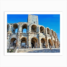 Roman Amphitheater in Arles. A majestic Roman amphitheater stands tall against a clear blue sky. The ancient structure is made of weathered stone, with numerous arches and a central tower. The amphitheater is surrounded by a set of stone steps leading up to the entrance. A few people are walking around the area, adding a sense of scale to the impressive monument. Art Print