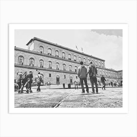 Exploring the Historic Building Palazzo Pitti. The image depicts a group of people walking and standing in front of a large, historic building with arched windows and a detailed facade. The scene is set in a spacious courtyard with a patterned stone pavement. The building's architecture and the presence of people suggest it is a place of interest, possibly a museum or a significant public building. Art Print