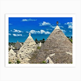 Trulli Houses in Italy. The image showcases a picturesque view of a traditional Italian village, featuring a cluster of Trulli houses with their distinctive conical roofs. The houses are constructed from white stone and have a rustic charm, with some roofs adorned with intricate patterns. The scene is bathed in warm sunlight, with a clear blue sky and fluffy white clouds overhead. 1 Art Print