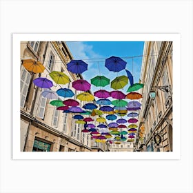 Colorful Umbrellas Street Art. A vibrant street scene features a multitude of colorful umbrellas suspended overhead, creating a whimsical and cheerful atmosphere. The umbrellas, in various shades of blue, green, purple, yellow, and pink, are strung across a narrow street lined with beige buildings. The buildings have white shutters and windows, adding to the European charm of the scene. Art Print