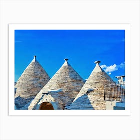 Trulli Houses in Italy. The image showcases three traditional Trulli houses, characterized by their distinctive conical roofs made of dry-stacked limestone. The houses are painted in a warm white, contrasting beautifully against the bright blue sky. The roofs are adorned with simple, white-painted finials, adding a touch of elegance. The scene is bathed in warm sunlight, casting soft shadows on the stone walls. Art Print