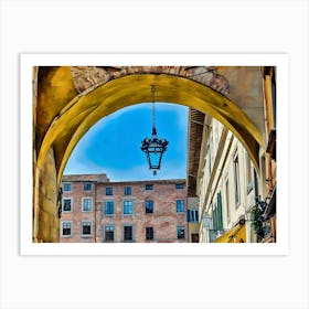 Lucca Under the Archway: A Glimpse of Historic Charm. This image captures a scenic view through a yellow-toned archway in a historic European city. A hanging lantern takes center stage, framed by rustic brick buildings and a bright blue sky, evoking the timeless allure of old-world architecture. Art Print