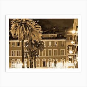 Nighttime scene with two palm trees in the foreground, their fronds silhouetted against a backdrop of a city street. The buildings are rendered in a sepia tone, with warm yellow lights emanating from windows and street lamps. The scene is bathed in a soft, warm glow, creating a sense of tranquility and charm. Art Print