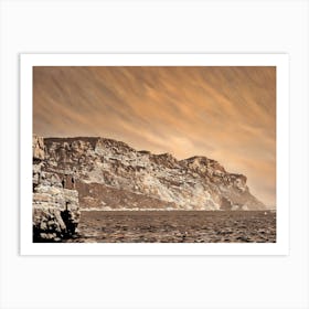 Cliffside View of Cap Canaille. The image captures a panoramic view of a rugged cliff face, its rocky surface bathed in warm, sepia tones. The cliff rises dramatically from the calm, blue waters below, creating a sense of grandeur and natural beauty. Two figures stand on a rocky outcrop at the edge of the cliff, their silhouettes barely visible against the vastness of the landscape. Art Print