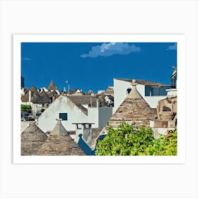 Trulli Rooftops in Italy. The image showcases a picturesque view of traditional Trulli houses in a small Italian town. The Trulli, characterized by their distinctive conical roofs made of dry-stone, are painted in a warm, earthy palette of browns and tans. The whitewashed walls of the houses contrast beautifully with the vibrant green foliage of a tree in the foreground. Art Print