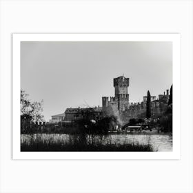 Medieval Castle by the Lake Garda Castello Scaligero Sirmione. A black and white photograph captures a majestic medieval castle standing tall against a cloudy sky. The castle, with its imposing stone walls and a prominent tower, dominates the scene. The foreground features a tranquil lake reflecting the castle's silhouette, with tall grasses and trees framing the view. Art Print