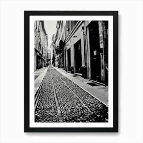 Cobblestone Alleyway in Pavia Italy. A black and white photograph captures a narrow alleyway lined with tall buildings. The path is paved with a cobblestone pattern, creating a textured surface. The buildings have closed doors and windows, suggesting a quiet and secluded atmosphere. The perspective is from the ground, looking down the alleyway, emphasizing the depth and narrowness of the space. Art Print