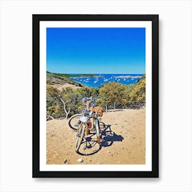 Bike Stop in Porquerolles with a Stunning Coastal View. Two bicycles rest on a sandy path overlooking a breathtaking Mediterranean bay dotted with anchored sailboats. The scene, framed by lush greenery and blue skies, captures the essence of a peaceful adventure along the coastline of Provence, France. Art Print