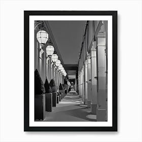 Paris Illuminated Corridor at Night. The image depicts a long, narrow corridor lined with large, geometric lanterns hanging from the ceiling. The corridor is flanked by tall, rectangular columns and features neatly trimmed, conical bushes in square planters along one side. The lighting creates a pattern of shadows on the floor, adding depth and perspective to the scene. Art Print