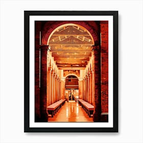 Brick Archway with Columns in Rimini historical center by night. The image depicts a narrow, arched passageway with a brick exterior. The passage leads into a long, colonnaded hall with a tiled floor. The hall is bathed in warm, orange light, creating a sense of depth and mystery. The ceiling is adorned with a complex pattern of wooden beams, adding to the architectural interest. Art Print