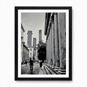 Towers and Columns in Pavia Italy. A black and white photograph captures a narrow street lined with tall, slender towers, their tops reaching towards a cloudy sky. The towers are made of stone and have a weathered, aged appearance. On the right side of the image, a row of imposing columns stands tall, creating a sense of grandeur and history. Art Print