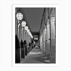 Paris Illuminated Corridor at Night. The image depicts a long, narrow corridor lined with large, geometric lanterns hanging from the ceiling. The corridor is flanked by tall, rectangular columns and features neatly trimmed, conical bushes in square planters along one side. The lighting creates a pattern of shadows on the floor, adding depth and perspective to the scene. Art Print