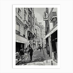 Paris Strolling Through a European Alley. The image depicts a charming European alleyway with cobblestone streets and classic architecture. People are seen walking along the narrow street, which is lined with quaint buildings, balconies, and various shop signs. The scene captures the essence of a leisurely stroll in a picturesque urban setting. Art Print