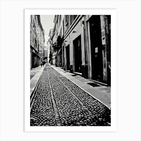 Cobblestone Alleyway in Pavia Italy. A black and white photograph captures a narrow alleyway lined with tall buildings. The path is paved with a cobblestone pattern, creating a textured surface. The buildings have closed doors and windows, suggesting a quiet and secluded atmosphere. The perspective is from the ground, looking down the alleyway, emphasizing the depth and narrowness of the space. Art Print