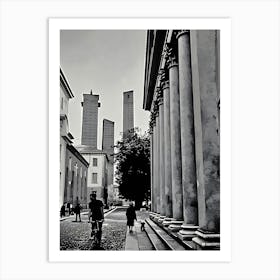 Towers and Columns in Pavia Italy. A black and white photograph captures a narrow street lined with tall, slender towers, their tops reaching towards a cloudy sky. The towers are made of stone and have a weathered, aged appearance. On the right side of the image, a row of imposing columns stands tall, creating a sense of grandeur and history. Art Print