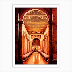 Brick Archway with Columns in Rimini historical center by night. The image depicts a narrow, arched passageway with a brick exterior. The passage leads into a long, colonnaded hall with a tiled floor. The hall is bathed in warm, orange light, creating a sense of depth and mystery. The ceiling is adorned with a complex pattern of wooden beams, adding to the architectural interest. Art Print