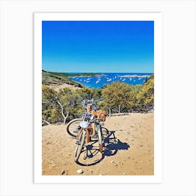 Bike Stop in Porquerolles with a Stunning Coastal View. Two bicycles rest on a sandy path overlooking a breathtaking Mediterranean bay dotted with anchored sailboats. The scene, framed by lush greenery and blue skies, captures the essence of a peaceful adventure along the coastline of Provence, France. Art Print