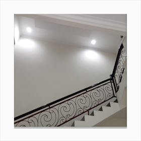 The staircase is characterized by its winding trails that lead up to the next level. The walls are painted in a pristine white color, which gives the space a modern and sleek feel. Soft lighting illuminates the stairs, providing a warm and welcoming atmosphere. Canvas Print