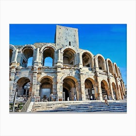 Roman Amphitheater in Arles. A majestic Roman amphitheater stands tall against a clear blue sky. The ancient structure is made of weathered stone, with numerous arches and a central tower. The amphitheater is surrounded by a set of stone steps leading up to the entrance. A few people are walking around the area, adding a sense of scale to the impressive monument. Canvas Print