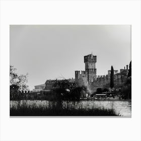 Medieval Castle by the Lake Garda Castello Scaligero Sirmione. A black and white photograph captures a majestic medieval castle standing tall against a cloudy sky. The castle, with its imposing stone walls and a prominent tower, dominates the scene. The foreground features a tranquil lake reflecting the castle's silhouette, with tall grasses and trees framing the view. Canvas Print