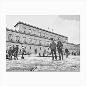 Exploring the Historic Building Palazzo Pitti. The image depicts a group of people walking and standing in front of a large, historic building with arched windows and a detailed facade. The scene is set in a spacious courtyard with a patterned stone pavement. The building's architecture and the presence of people suggest it is a place of interest, possibly a museum or a significant public building. Canvas Print