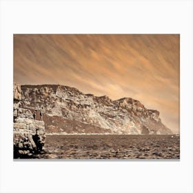 Cliffside View of Cap Canaille. The image captures a panoramic view of a rugged cliff face, its rocky surface bathed in warm, sepia tones. The cliff rises dramatically from the calm, blue waters below, creating a sense of grandeur and natural beauty. Two figures stand on a rocky outcrop at the edge of the cliff, their silhouettes barely visible against the vastness of the landscape. Canvas Print