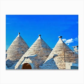 Trulli Houses in Italy. The image showcases three traditional Trulli houses, characterized by their distinctive conical roofs made of dry-stacked limestone. The houses are painted in a warm white, contrasting beautifully against the bright blue sky. The roofs are adorned with simple, white-painted finials, adding a touch of elegance. The scene is bathed in warm sunlight, casting soft shadows on the stone walls. Canvas Print