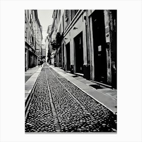 Cobblestone Alleyway in Pavia Italy. A black and white photograph captures a narrow alleyway lined with tall buildings. The path is paved with a cobblestone pattern, creating a textured surface. The buildings have closed doors and windows, suggesting a quiet and secluded atmosphere. The perspective is from the ground, looking down the alleyway, emphasizing the depth and narrowness of the space. Canvas Print