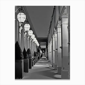 Paris Illuminated Corridor at Night. The image depicts a long, narrow corridor lined with large, geometric lanterns hanging from the ceiling. The corridor is flanked by tall, rectangular columns and features neatly trimmed, conical bushes in square planters along one side. The lighting creates a pattern of shadows on the floor, adding depth and perspective to the scene. Canvas Print