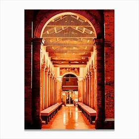 Brick Archway with Columns in Rimini historical center by night. The image depicts a narrow, arched passageway with a brick exterior. The passage leads into a long, colonnaded hall with a tiled floor. The hall is bathed in warm, orange light, creating a sense of depth and mystery. The ceiling is adorned with a complex pattern of wooden beams, adding to the architectural interest. Canvas Print